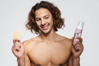 Beyaz arka planda izole edilmiş kozmetik sünger ve temizlik köpüğüyle poz veren mutlu üstsüz adam.