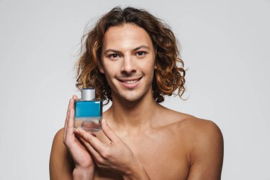 Beyaz arka planda izole edilmiş parfümle poz verirken gülümseyen tişörtsüz adam.