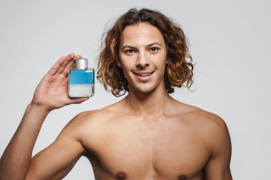 Beyaz arka planda izole edilmiş parfümle poz verirken gülümseyen tişörtsüz adam.