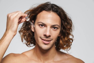 Beyaz arka planda izole edilmiş yüz serumu uygularken gülümseyen tişörtsüz adam.