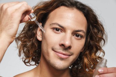 Beyaz arka planda izole edilmiş yüz serumu uygularken gülümseyen tişörtsüz adam.