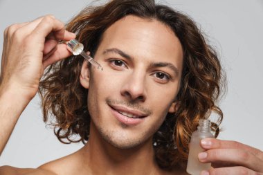 Beyaz arka planda izole edilmiş yüz serumu uygularken gülümseyen tişörtsüz adam.