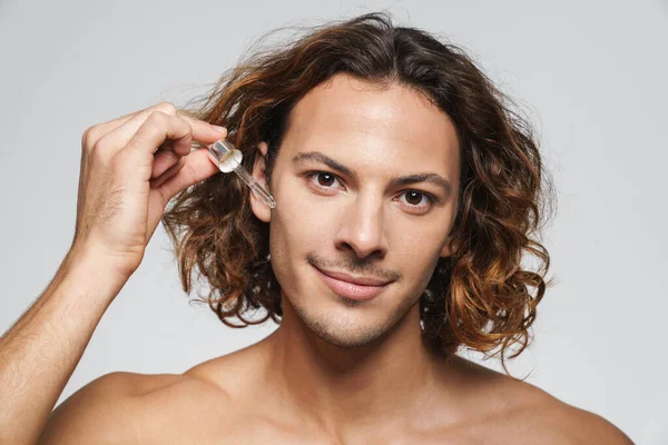 Beyaz arka planda izole edilmiş yüz serumu uygularken gülümseyen tişörtsüz adam.