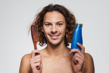 Tarak ve şampuanla poz veren mutlu tişörtsüz adam beyaz arka planda izole edilmiş.