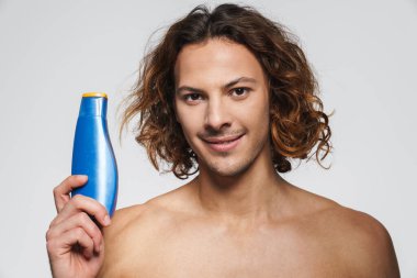 Beyaz arka planda izole edilmiş şampuanla poz verirken gülümseyen tişörtsüz adam.
