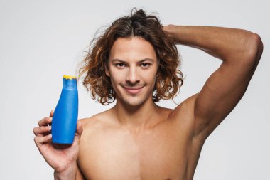 Beyaz arka planda izole edilmiş şampuanla poz verirken gülümseyen tişörtsüz adam.