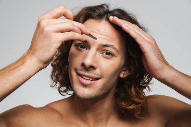 Beyaz arka planda izole edilmiş cımbızla kaşlarını yolarken gülümseyen mutlu gömleksiz adam.