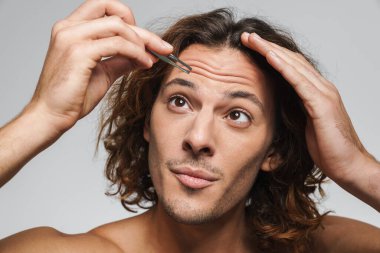 Beyaz arka planda izole edilmiş cımbızla kaşlarını koparan odaklanmış, yakışıklı bir adam.