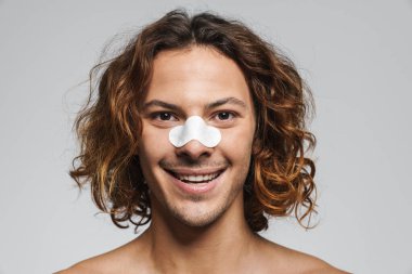 Beyaz duvardan izole edilmiş siyah başlıkları çıkarmak için burnunda temiz yamalarla poz veren neşeli gömleksiz adam.