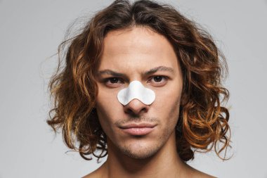 Beyaz duvardan izole edilmiş siyah başlıkları çıkarmak için burnunda temiz yamalar olan ciddi tişörtsüz bir adam.