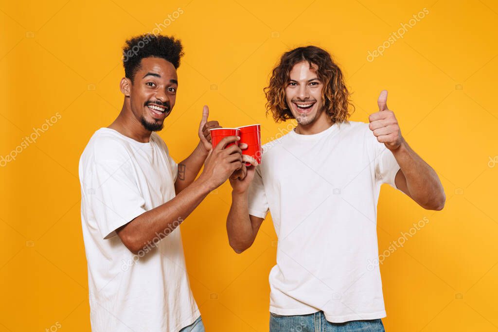 Two happy male friends celebrating, holding plastic cups isolated over yellow background, cheering