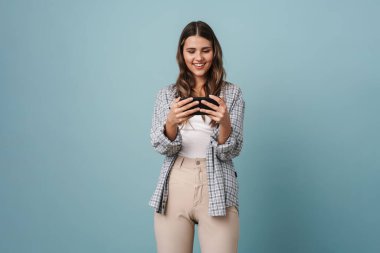 Mavi arka planda izole edilmiş cep telefonuyla online oyun oynayan neşeli güzel kız.
