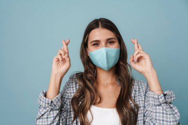 Yüzünde maske olan mutlu bir kız şans dilemek için mavi arka planda poz veriyor.