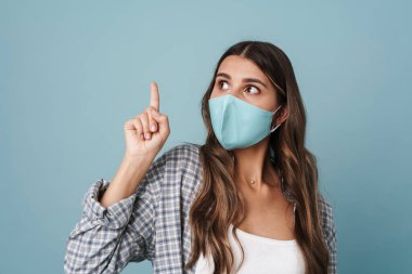 Yüz maskesi takmış şaşırmış bir kız yana bakıyor ve mavi arka planda yalnız bir şekilde parmağını yukarı kaldırıyor.