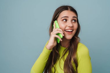 Mavi arka planda izole bir şekilde cep telefonuyla konuşurken gülümseyen neşeli, güzel kız.