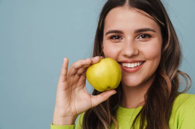 Gülümseyen ve elma gösteren neşeli güzel kız mavi arka planda izole edilmiş.