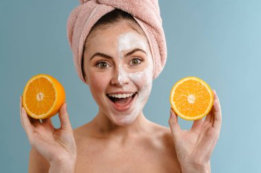 Kafasına banyo havlusu takan, portakal gösteren, kozmetik maske takan güzel genç bir kadın.