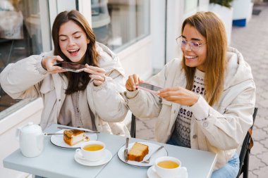 Genç neşeli kız arkadaşlar kafede oturup cep telefonuyla yemeklerin fotoğrafını çekiyorlar.