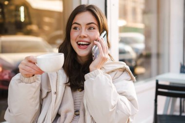 Kahve dükkanında otururken cep telefonu kullanan güzel, gülümseyen bir kadın. Elinde fincanla.