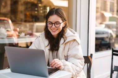 Açık kafede otururken dizüstü bilgisayar kullanan gülümseyen genç kadın.