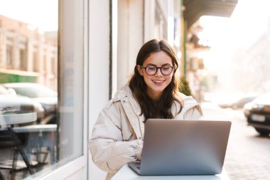 Açık kafede otururken dizüstü bilgisayar kullanan gülümseyen genç kadın.
