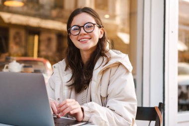 Açık kafede otururken dizüstü bilgisayar kullanan gülümseyen genç kadın.