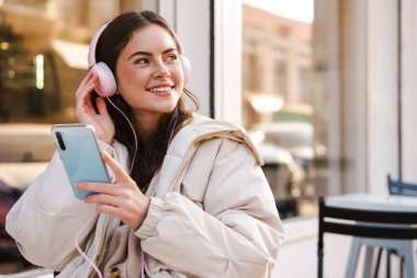 Genç bir kadın kahve dükkanında oturmuş, müzik dinliyor ve elinde akıllı bir telefonla mesaj atıyor.