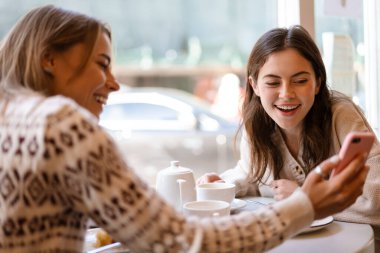 Kafede öğle yemeği yerken cep telefonu kullanan iki gülümseyen genç kız arkadaş.