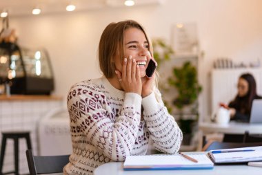 Kapalı kafede ödev yaparken cep telefonuyla konuşan sevimli öğrenci kız.
