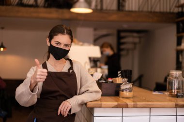 Medikal koruyucu maskeli memnun garson kafedeki kamerada baş parmağını gösteriyor.