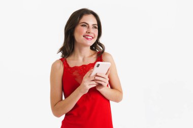 Kırmızı elbise giyen neşeli güzel kız beyaz arka planda izole edilmiş cep telefonu kullanıyor.
