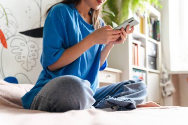 Gülümseyen genç bir kadın evde yatağında otururken cep telefonu kullanıyor. Kesik bir görüntü.