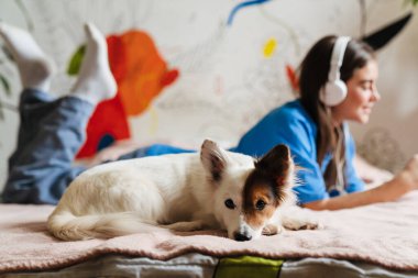 Kulaklıklı neşeli kız evde köpeğiyle yatakta yatıyor.