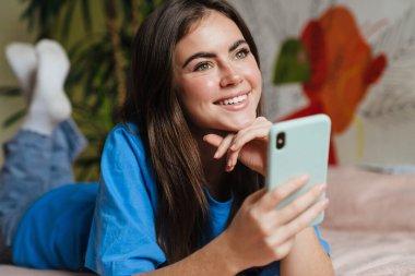 Mutlu esmer güzel kız evde yatarken cep telefonu kullanıyor.