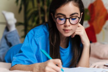 Gözlüklü esmer kız evde yatakta yatarken ödev yapıyor.