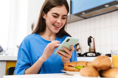 Mutfakta sandviç yerken cep telefonu kullanan mutlu esmer kız.