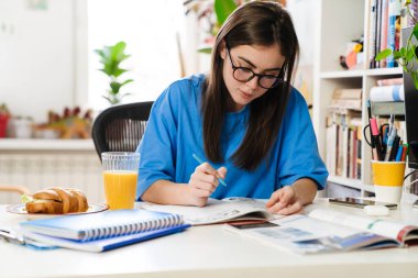 Gözlüklü esmer kız oturma odasında masada otururken ödev yapıyor.