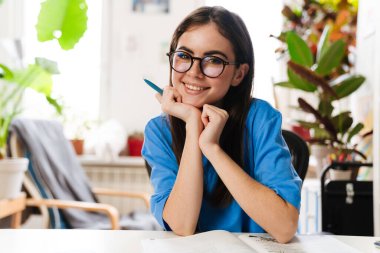 Gözlüklü mutlu esmer kız ev ödevi yapıyor ve evde gülümsüyor.