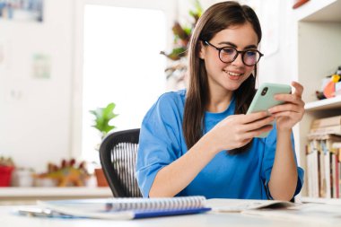 Gözlüklü mutlu esmer kız evde ödev yaparken akıllı telefon kullanıyor.