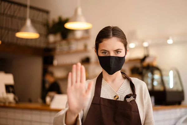 Serious young barista forbid customers come inside coffee shop without medical mask, showing stop gesture