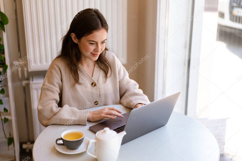 Smiling charming girl working with laptop while drinking tea in cafe indoors