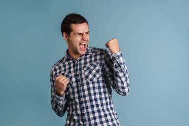 Heyecanlı genç adam çığlık atarken kazanan jest yapıyor mavi arka planda izole edilmiş