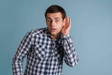 Şok olmuş genç esmer adam yana bakıyor ve mavi arka planda izole edilmiş kulak misafiri.