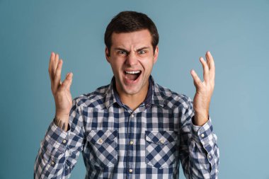 Kızgın genç adam mavi arka planda izole bir şekilde çığlık atıyor.