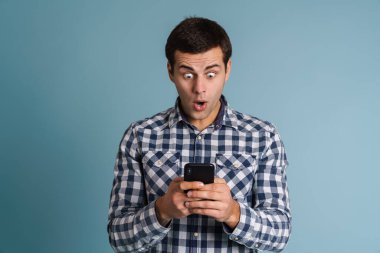 Mavi arka planda izole edilmiş cep telefonu tutan kafası karışmış genç bir adamın portresi.