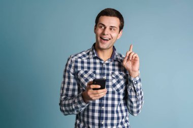 Ekoseli tişört giyen, elinde cep telefonu tutan ve mavi arka planda izole edilmiş bir fikri olan genç adam.