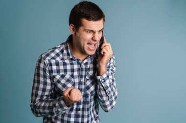 Mavi arka planda izole bir şekilde cep telefonu kullanan ve bağıran öfkeli bir adamın portresi.