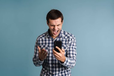 Mavi arka planda izole bir şekilde cep telefonu kullanan ve bağıran öfkeli bir adamın portresi.