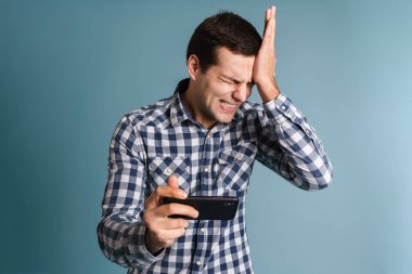 Kafası karışmış genç adam mavi arka plan yerine akıllı telefon kullanıyor, oyunlar oynuyor.