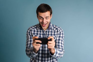 Heyecanlı genç adam mavi arka plan yerine akıllı telefon kullanıyor, oyunlar oynuyor.